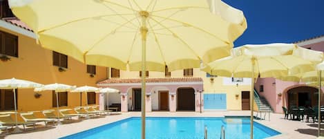 Un espace piscine agrémenté de parasols jaunes, de chaises longues et d’un bâtiment doté d’un toit en tuiles.