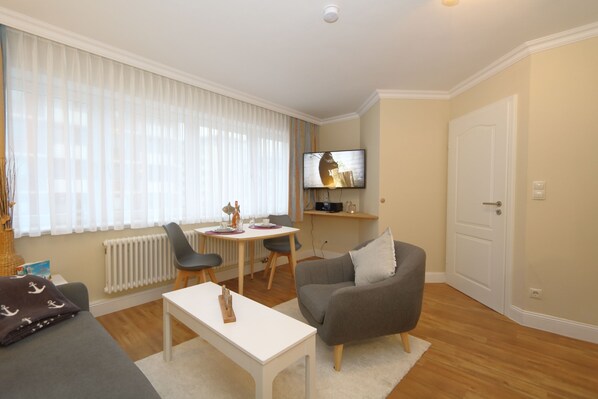 A living room with a sofa, a coffee table, a TV mounted on the wall, a dining table with chairs, and a radiator.