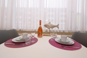 A dining table set with plates, glasses, cutlery, and a bottle of liquor, with a decorative fish ornament and sheer curtains in the background. - Haus Sarah 04, Deutschland (Sylt)