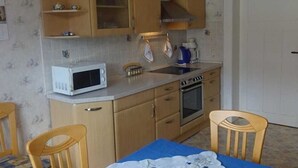 A compact kitchen with wooden cabinets, a microwave, a sink, and an oven. There is a dining table with chairs and a blue tablecloth. - Ferienwohnung Herrmann (Weischlitz)