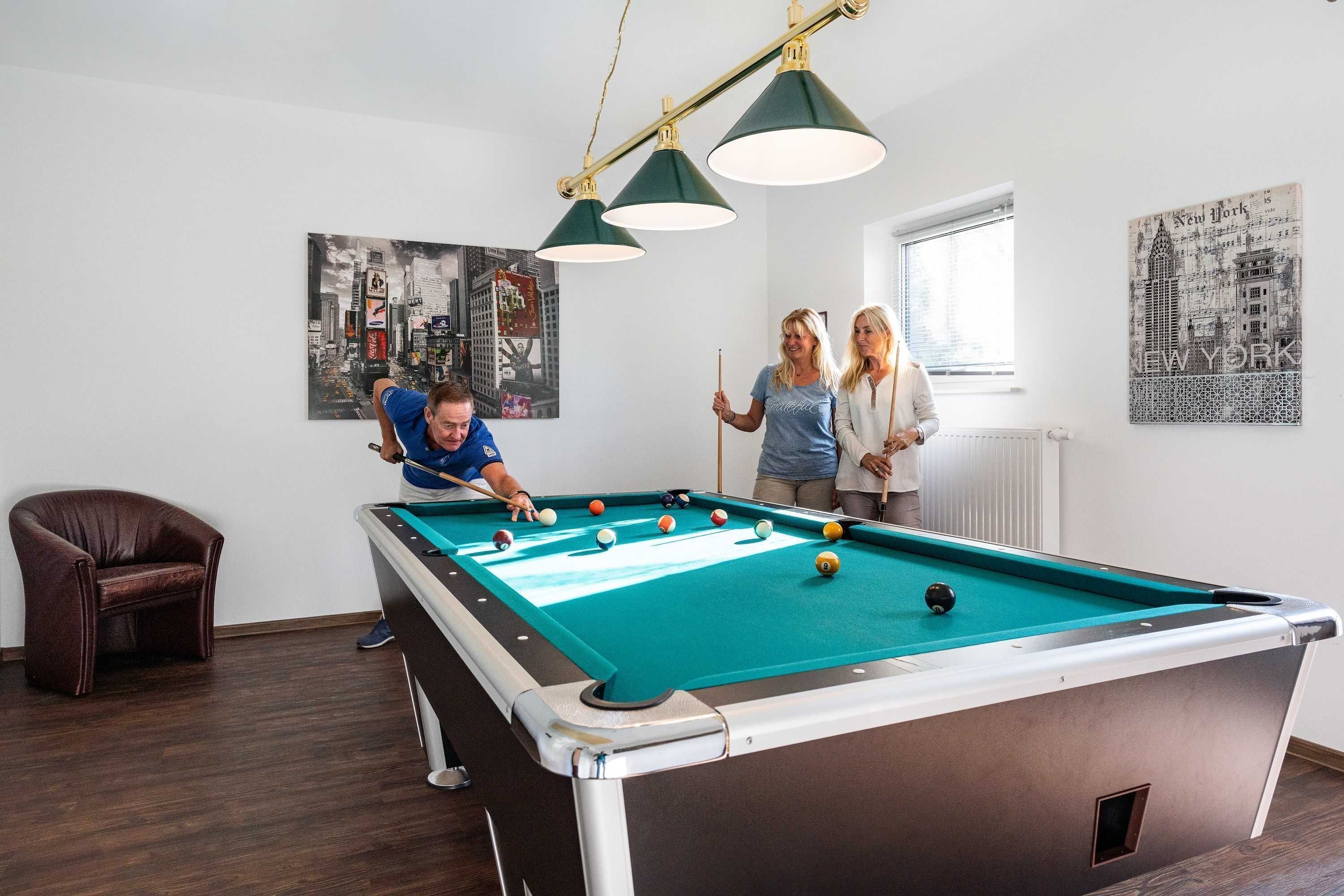 Ett biljardbord i ett rum med en brun läderfåtölj, en målning som föreställer en stadsbild och en affisch med motiv från New York på väggen.