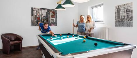 Ett biljardbord i ett rum med en brun läderfåtölj, en målning som föreställer en stadsbild och en affisch med motiv från New York på väggen.