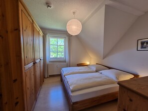 A bedroom with a wooden wardrobe, a window with a view, a bed with pillows, and a hanging light fixture.