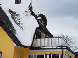 A yellow house with a steep roof covered in snow, a white balcony, and a window.