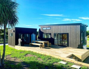 A modern, two-story house with a deck, large windows, and a covered outdoor seating area. - Oasis in the Heads (Mangawhai)