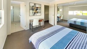 A bedroom with a bed, a desk, and a chair. There is a mirror on the wall and a ceiling fan. - Lillypilly Resort Apartments (Gracemere)