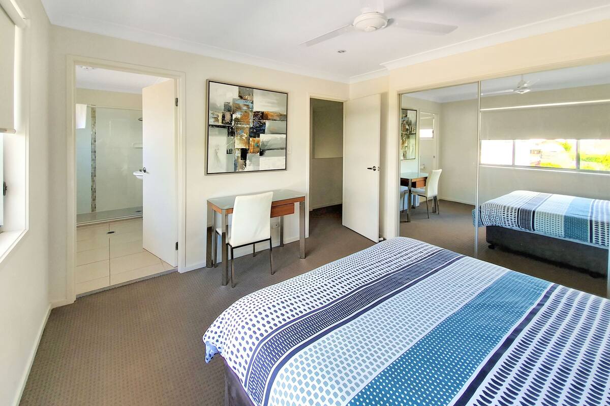 A bedroom with a bed, a desk, and a chair. There is a mirror on the wall and a ceiling fan.