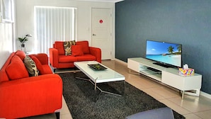 A modern living room with a red sofa, a white coffee table, and a flat-screen TV on a white TV stand. - Lillypilly Resort Apartments (Gracemere)