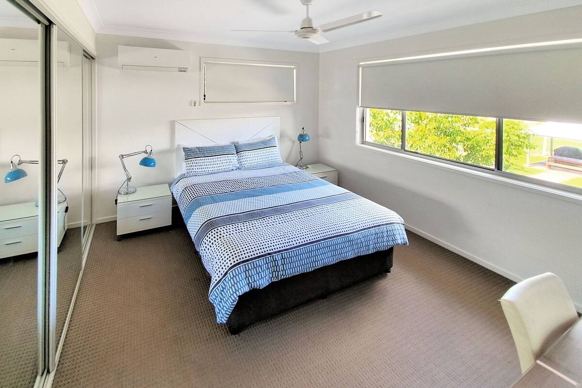 A bedroom with a bed, bedside tables, a ceiling fan, a window with a view, and a mirror.