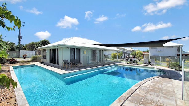 A rectangular swimming pool with a tiled deck, a covered seating area with chairs, and a clear blue sky. - Lillypilly Resort Apartments (Gracemere)