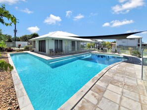 A rectangular swimming pool with a tiled deck, a covered seating area with chairs, and a clear blue sky. - Lillypilly Resort Apartments (Gracemere)