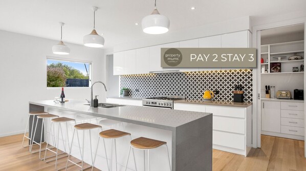 A modern kitchen with a central island, bar stools, pendant lights, and a 'PAY 2 STAY 3' sign. - Macks Family Hideout (Safety Beach)