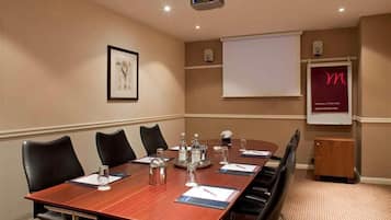 Un salón de conferencias con una larga mesa, sillas, un proyector, una pizarra y un cuadro en la pared.