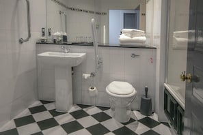 Baño con lavamanos blanco, inodoro y ducha. Hay toallas apiladas en el mostrador y un escobilla para el inodoro.