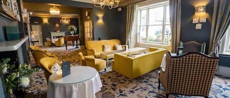 A hotel room with a fireplace, a dining table, yellow armchairs, a patterned carpet, and a large window.