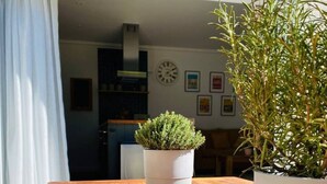 Ein Holztisch mit einer Topfpflanze in der Mitte, ein weißer Vorhang und eine Küche im Hintergrund.