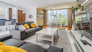 A modern living room with a black leather sofa, a white coffee table, and a TV on a white stand. There is a balcony with a view of the city.