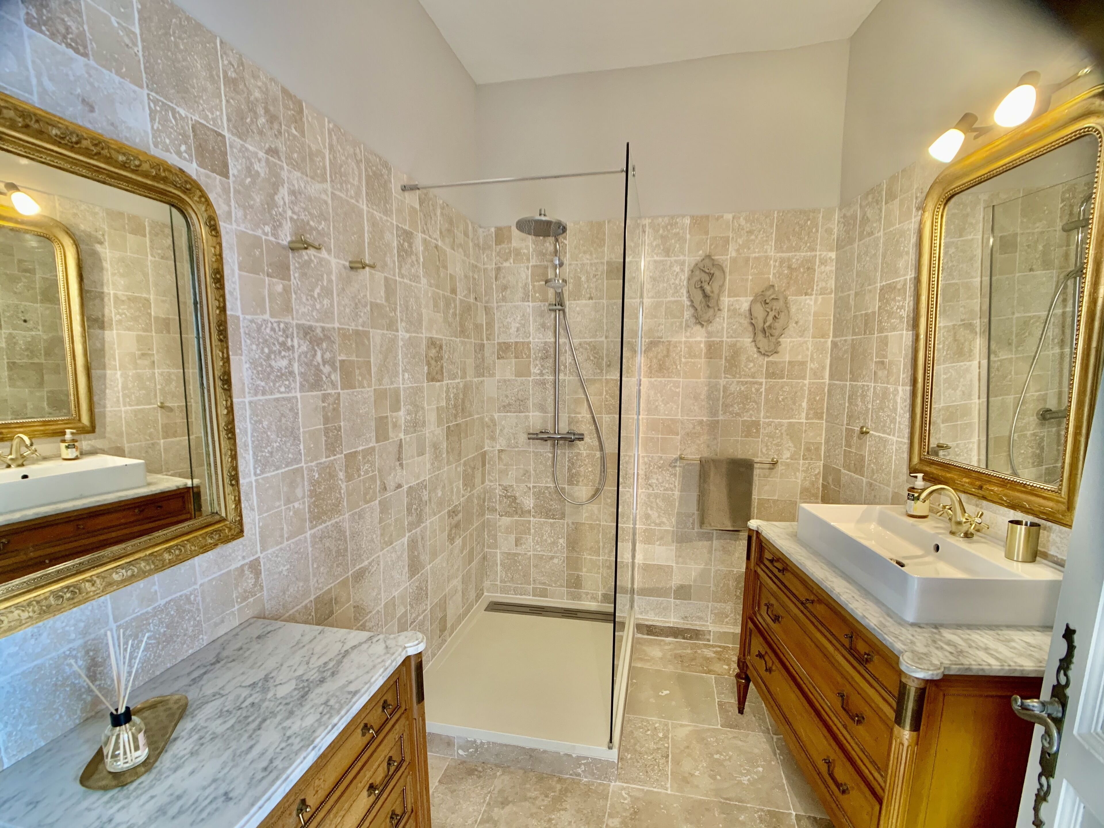 A bathroom with two sinks, a glass shower enclosure, marble countertops, and ornate gold-framed mirrors.