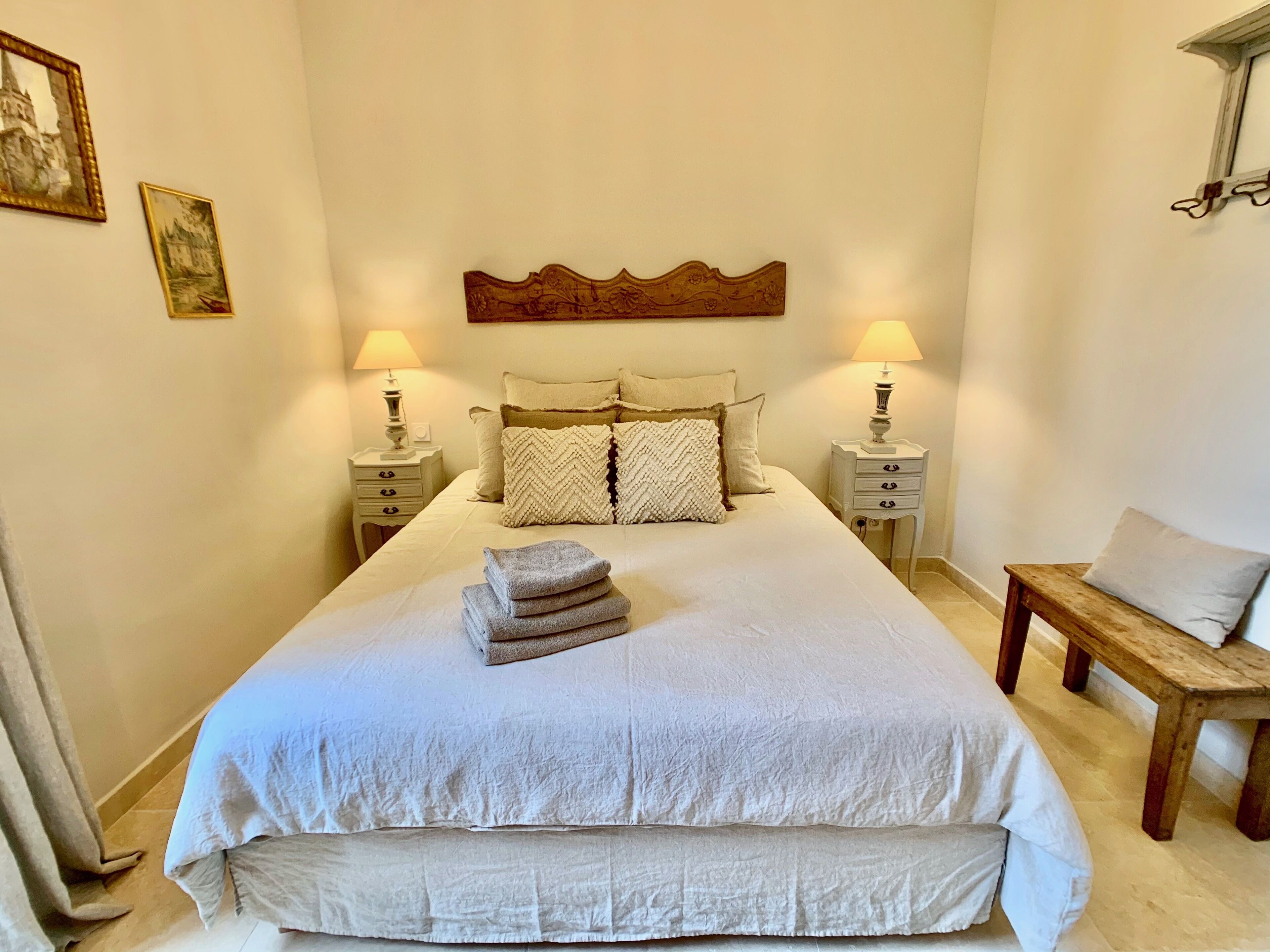Ein ordentlich bezogenes Bett mit Kopfteil, zwei Nachttische mit Lampen, eine hölzerne Bank und ein an der Wand befestigter Bilderrahmen.
