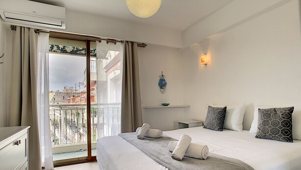A hotel room with a bed, a desk, a chair, an air conditioner, a window with curtains, and a view of buildings. - N&J - SUNRISE TERRACE - Close sea - Terrace 30m² (Niza)