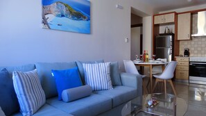 A modern living room with a sofa, a dining table, and chairs. There is a kitchen area with cabinets and appliances. A painting is on the wall.