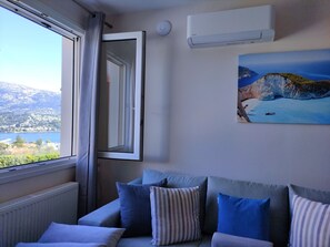 A room with a view of a mountainous landscape through a window, a white air conditioning unit, and a painting of a coastal scene.