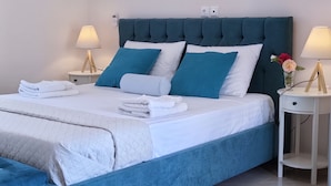 Una cama bien hecha con ropa de cama azul y blanca, dos mesitas de noche con lámparas, y un jarrón con flores sobre una de ellas.