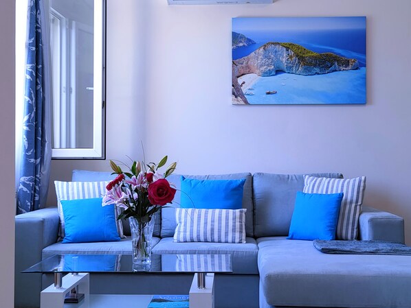 A modern living room with a grey sofa, blue cushions, a glass coffee table, and a vase with flowers.