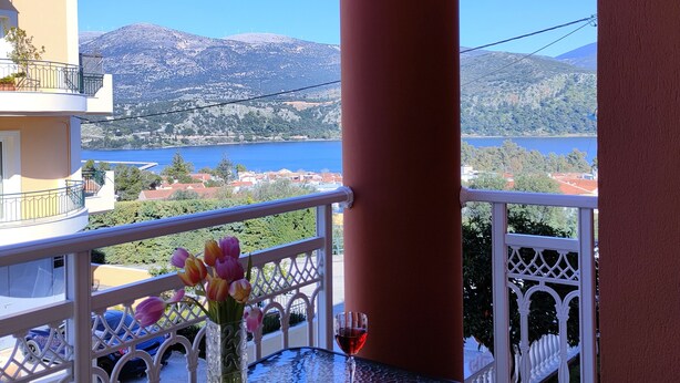 Un balcón con una mesa dispuesta para dos personas, que incluye una copa de vino tinto y un jarrón con flores. El balcón ofrece vistas panorámicas de un lago y montañas.