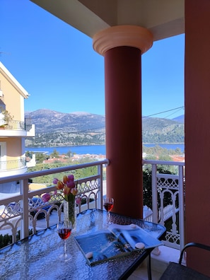 A balcony with a table set for two, featuring a glass of red wine and a vase with flowers. The balcony overlooks a scenic view of a lake and mountains.