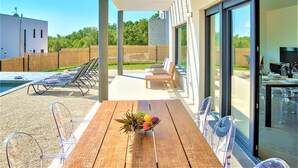 Ein Holztisch mit Obstschale, transparente Kunststoffstühle, ein Pool und eine überdachte Terrasse mit Blick auf die Grünanlage.