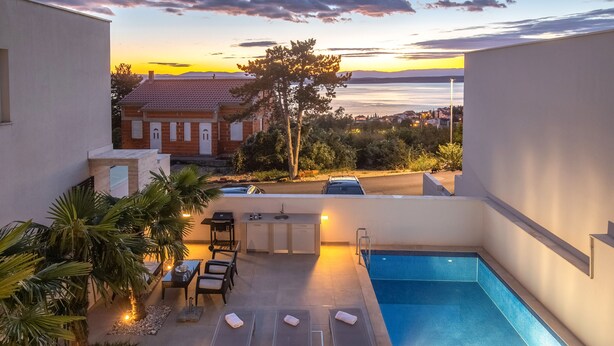 Un espace piscine aménagé sur le toit, avec des chaises longues et une table à manger, offrant une vue sur le paysage urbain au coucher du soleil.