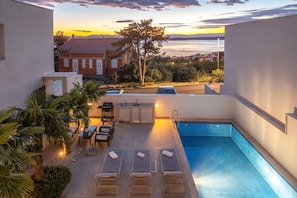 A rooftop pool area with lounge chairs and a dining table, overlooking a cityscape at sunset.