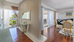 Un vaste espace de vie comprenant une table basse en verre, un canapé et une table à manger avec des chaises. Un grand miroir orne le mur et une porte-fenêtre coulissante offre une vue sur l’extérieur.