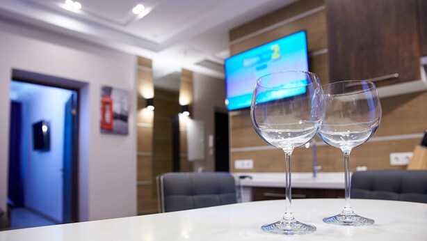 Una habitación de hotel moderna con una mesa blanca, dos copas de vino vacías, un televisor en la pared y una puerta que da a otra habitación.