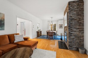 Una sala de estar con un sofá marrón, un comedor con mesa y sillas, una chimenea y un pasillo con una puerta.