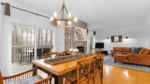 A spacious living room with a wooden dining table, comfortable seating, a fireplace, and a large window overlooking a forested area.