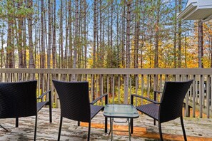 Una terraza de madera con dos sillas de mimbre y una mesita pequeña, con vistas a un bosque de árboles altos.