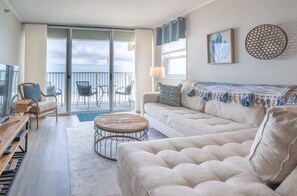 Ein geräumiges Wohnzimmer mit einer Couch, einem runden Couchtisch und einem Fernseher. Es gibt einen Balkon mit Stühlen und einem Tisch.