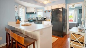 A kitchen with white cabinets, a black refrigerator, a dining area with wooden chairs, and a ceiling fan. - Salty Hammocks B1 (Carolina Beach)