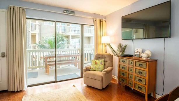 A room with a sliding glass door leading to a balcony, a beige armchair, a wooden dresser with drawers, a flat-screen TV mounted on the wall, and a lamp on a side table. - Salty Hammocks B1 (Carolina Beach)