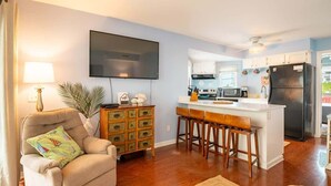 A living room with a sofa, a TV, a wooden cabinet, and a kitchen area with a refrigerator, stove, and bar stools. - Salty Hammocks B1 (Carolina Beach)