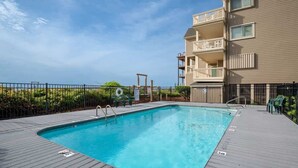 A swimming pool with a wooden deck, surrounded by a fence, and adjacent to a building with balconies. - Salty Hammocks B1 (Carolina Beach)