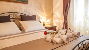 A neatly made bed with rolled towels and a rose, a bedside table with a lamp, and a painting on the wall.