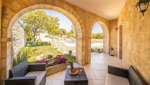 A covered outdoor area with stone arches, wicker furniture, and a view of a pool and garden.