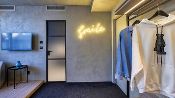 Una habitación moderna con un letrero de neón que dice 'Smile', un televisor, un perchero con ropa y una pequeña mesa con un florero.