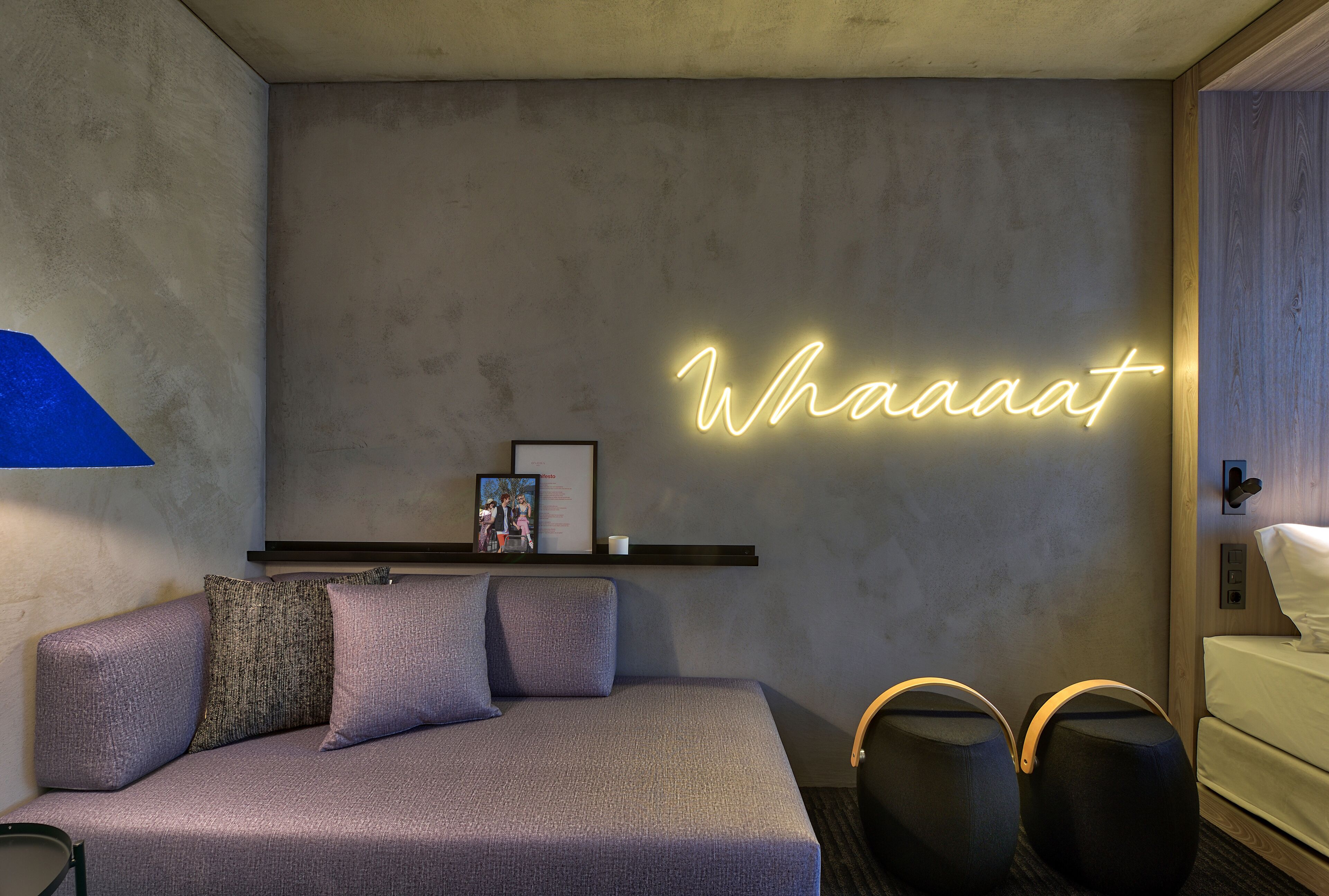 Una camera d'albergo moderna con un divano grigio, un neon con la scritta "Whaaat", un piccolo ripiano con una cornice e due pouf neri.