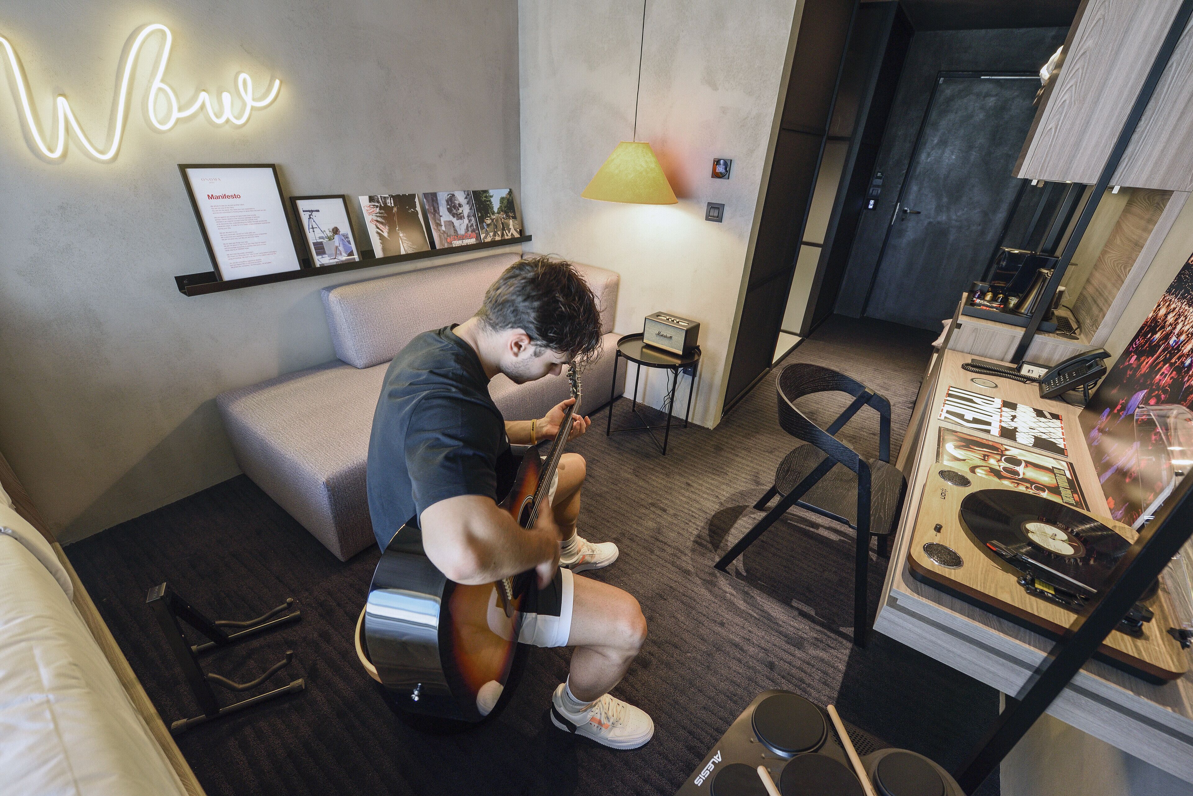 Une pièce avec un lit, une guitare, un bureau avec un ordinateur et un mur orné d’un néon.