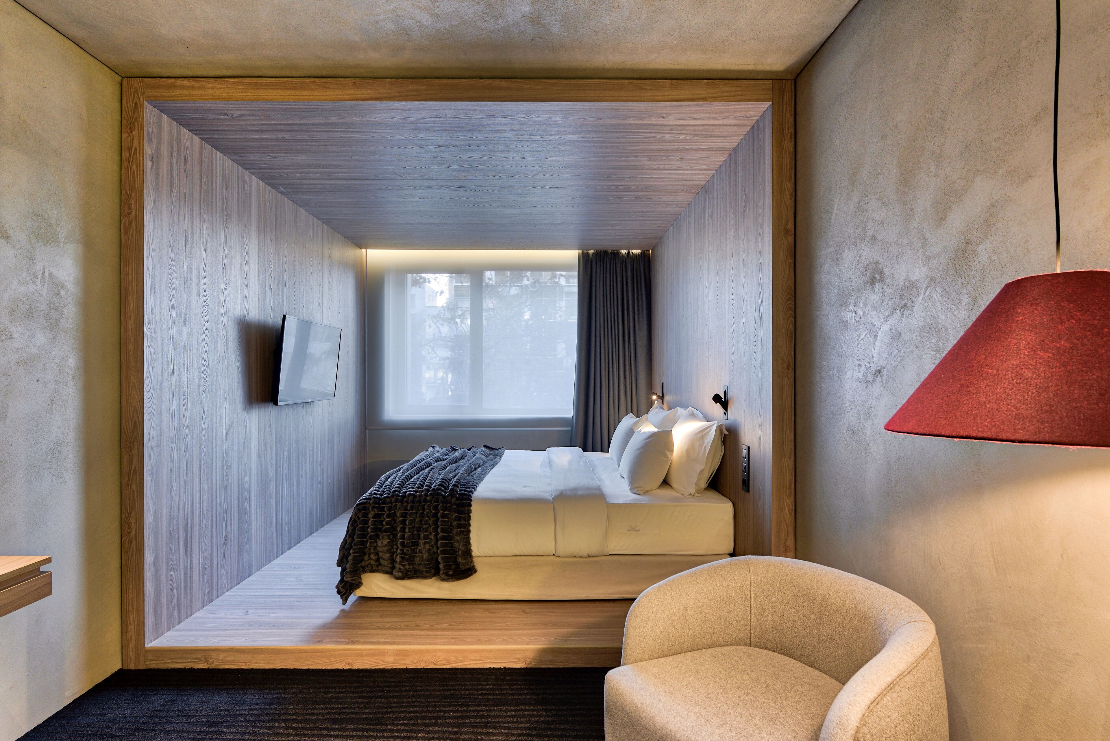Quarto compacto com cama, uma cadeira e uma janela.