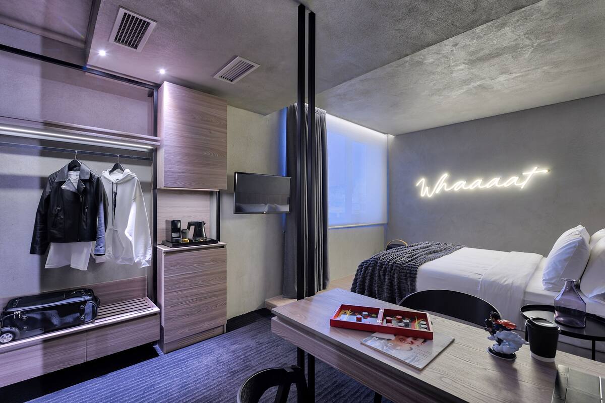 A modern hotel room with a bed, a desk with a laptop, a TV, a wardrobe, and a neon sign.